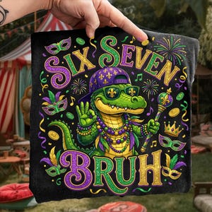 Könnte beinhalten: Schwarzes grafisches Handtuch mit einem grünen Alligator, der einen lila Hut und eine Sonnenbrille trägt und ein Zepter hält. Auf dem Handtuch steht "SIX SEVEN BRUH" in Gold, Grün und Lila. Mardi Gras-Thema mit Masken, Noten und Münzen.