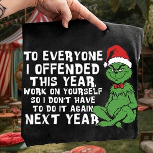 Op de afbeelding: Zwart kussen met de Grinch met een kerstmuts en rode vlinderdas. De tekst luidt: "TO EVERYONE I OFFENDED THIS YEAR, WORK ON YOURSELF SO I DON'T HAVE TO DO IT AGAIN NEXT YEAR."