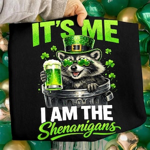 May include: Black graphic tee featuring a raccoon wearing a green hat and shamrock glasses, holding a green beer. Text reads "IT'S ME" above and "I AM THE Shenanigans" below the raccoon illustration. The design is perfect for St. Patrick's Day.