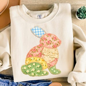 May include: Cream-colored sweatshirt featuring a patchwork bunny design. The bunny is composed of floral, gingham, and lace fabrics in pink, yellow, and green. The sweatshirt has a crew neck and long sleeves, perfect for spring.