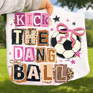 May include: White tote bag with the text "KICK THE DANG BALL" in various fonts and colors. A soccer ball with a pink bow and star accents is also featured. The bag has a playful, sporty design.
