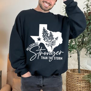 May include: Black crew neck sweatshirt featuring a white graphic of the Texas state outline, a star, and a floral design. The text reads "Hill Country Stronger Than The Storm".