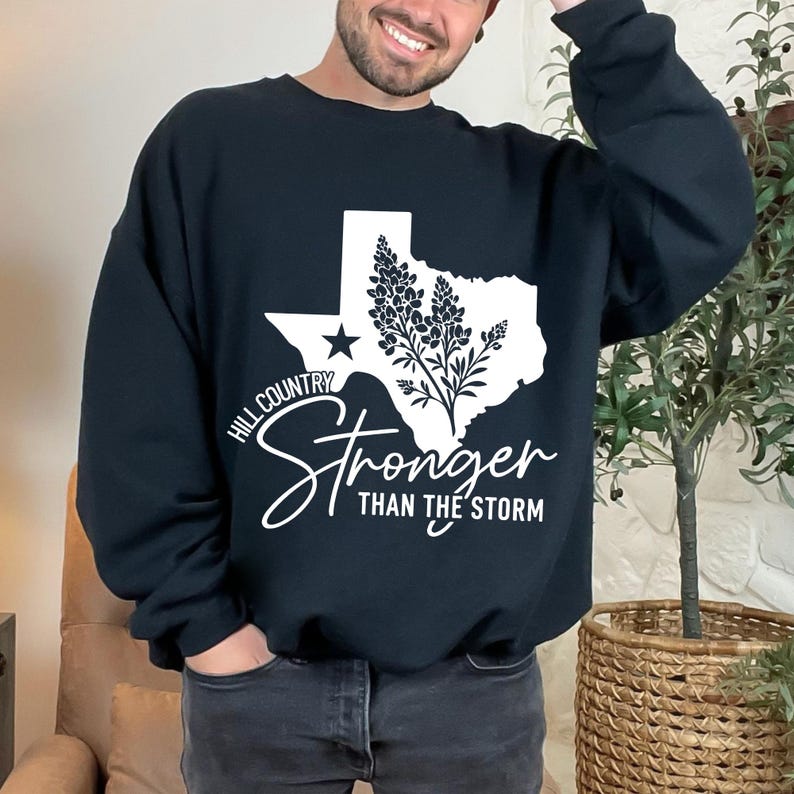 May include: Black crew neck sweatshirt featuring a white graphic of the Texas state outline, a star, and a floral design. The text reads "Hill Country Stronger Than The Storm".