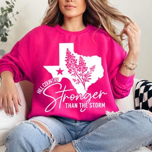 May include: A vibrant pink sweatshirt with a white graphic of the Texas state outline, a star, and floral elements. The text "Hill Country Stronger Than the Storm" is printed in white.