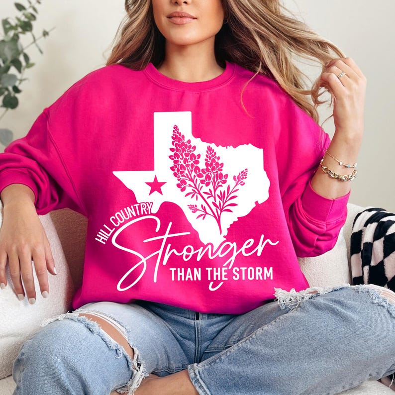 May include: A vibrant pink sweatshirt with a white graphic of the Texas state outline, a star, and floral elements. The text "Hill Country Stronger Than the Storm" is printed in white.