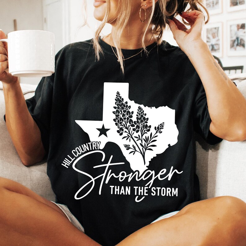 May include: Black t-shirt with a white graphic of the Texas state outline, a star, and floral elements. The shirt displays the text "Hill Country Stronger Than The Storm" in a cursive font.