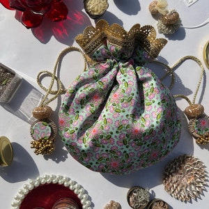 Könnte beinhalten: Eine grüne Kordeltasche mit Blumenmuster und goldenen Verzierungen. Die Tasche ist mit goldenen Quasten und Perlen verziert. Die Tasche ist von verschiedenen Dekorationsartikeln umgeben, darunter eine rote Lotusblume, eine kleine Schüssel Reis und ein kleines Glas Glitzer.