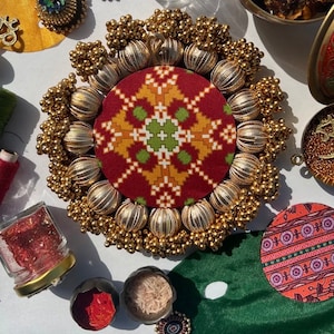 May include: A round, gold-coloured decorative piece with a red and gold fabric centre. The fabric has a geometric pattern. The piece is surrounded by small, gold beads.