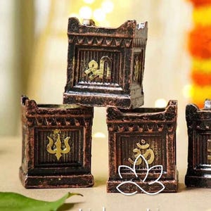 May include: Bronze-coloured decorative boxes with gold religious symbols. The boxes are stacked and arranged in a group. The text "Urban Utsav" is at the bottom of the image.