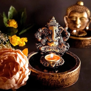 May include: A gold-toned metal incense burner with a small, lit candle in the center. A gold-toned metal statue of Ganesha sits on top of the burner. The burner is on a dark surface.