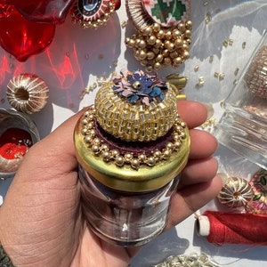Op de afbeelding: Een klein glazen potje met een decoratief goudkleurig deksel, vastgehouden in een hand. Het deksel is versierd met kleine kralen en een bloemmotief. Andere decoratieve elementen, waaronder rode glazen stukken en kralenornamenten, zijn zichtbaar.
