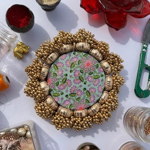 May include: A circular decorative piece with a gold beaded border and a floral patterned centre. The centre is a light green fabric with pink flowers. The piece is surrounded by various small containers with beads and other decorative items.