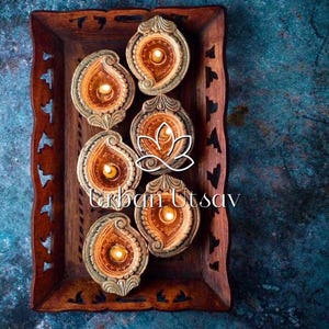 Op de afbeelding: Zes sierlijke, goudkleurige, traanvormige diya-lampen met brandende lonten, gerangschikt in een houten dienblad met ingewikkelde houtsnijwerk. De diya-lampen zijn een traditioneel Indiaas symbool voor licht en hoop.