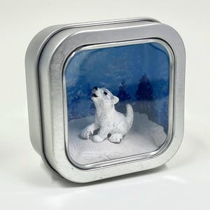 May include: A small, silver tin with a clear window displaying a miniature winter scene. A white wolf figurine is howling in a snowy landscape with blue trees and sky. The tin is square with rounded edges.