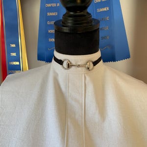 May include: A black choker necklace with a silver horse bit pendant. The necklace is on a white mannequin with a blue ribbon in the background.