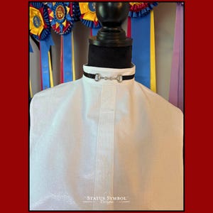 May include: A white equestrian show shirt with a black collar featuring silver-toned bit hardware. The shirt is displayed on a mannequin with a black neck form. The background includes colorful ribbons.