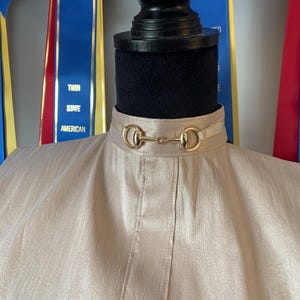 May include: A beige show shirt with a gold snaffle bit collar. The shirt is on a mannequin with blue and red ribbons behind it. The ribbons have the text "Twin State American" on them.