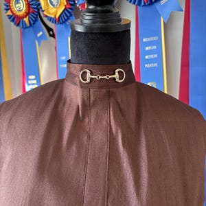 May include: A brown, high-collared garment with a decorative gold-colored bit closure. The fabric has a subtle sheen. The background includes blue ribbons with text, suggesting a competition or event.