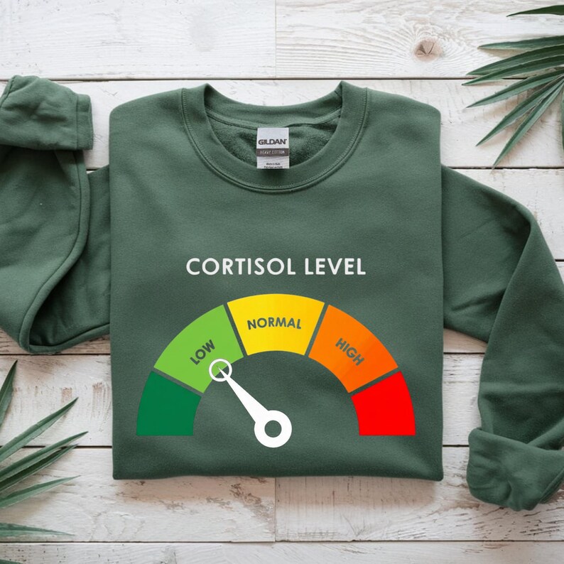 Puede incluir: Sudadera verde oscuro con el texto "CORTISOL LEVEL" sobre un indicador. El indicador muestra "LOW", "NORMAL" y "HIGH" en verde, amarillo y rojo. Una aguja blanca se&ntilde;ala el nivel "NORMAL".