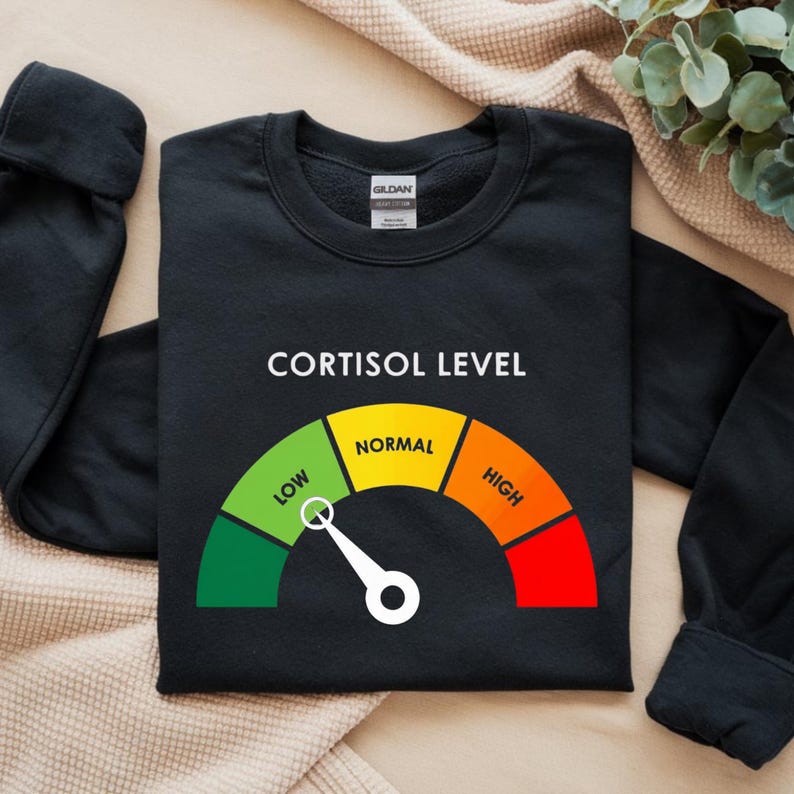 Puede incluir: Sudadera negra con la frase "CORTISOL LEVEL" y un gr&aacute;fico de indicador. El indicador muestra "BAJO" en verde, "NORMAL" en amarillo y "ALTO" en rojo. La aguja apunta hacia "NORMAL". La sudadera es de material suave.