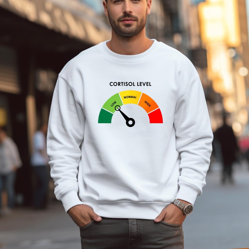 Puede incluir: Sudadera blanca con un gr&aacute;fico de un medidor de nivel de cortisol. El medidor muestra los niveles "Bajo", "Normal" y "Alto" en verde, amarillo y rojo. El texto "CORTISOL LEVEL" est&aacute; encima del medidor. La sudadera la lleva una persona.