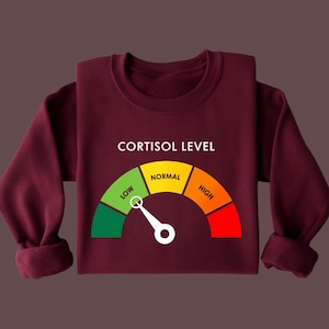 Puede incluir: Una sudadera burdeos con la frase "CORTISOL LEVEL" en blanco. Debajo, un indicador con una aguja que se&ntilde;ala "NORMAL". El indicador tiene secciones verdes, amarillas, naranjas y rojas etiquetadas como "LOW" y "HIGH".