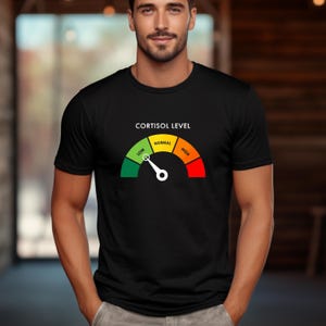 Puede incluir: Camiseta negra con un gr&aacute;fico de medidor de nivel de cortisol. El medidor muestra un espectro de verde (bajo) a rojo (alto), con "Normal" en amarillo. El texto "CORTISOL LEVEL" est&aacute; encima del medidor. Una camiseta casual.