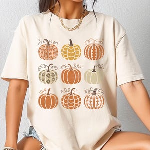 May include: A cream-colored short-sleeve t-shirt featuring a graphic print of nine pumpkins arranged in a three-by-three grid. Each pumpkin displays a unique pattern, including swirls, stripes, dots, and floral motifs in shades of orange, tan, and white.