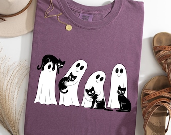 May include: A plum-colored t-shirt featuring a graphic of four white ghosts interacting with black cats. The design shows ghosts holding or sitting with the cats. The shirt has a crew neckline and a Comfort Colors brand label on the inner collar.