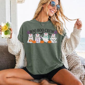 May include: A sage green t-shirt featuring a graphic of four ghost nurses wearing nurse caps and stethoscopes, with the text "BOO BOO CREW" printed above them. The design includes small orange pumpkins, purple spiderwebs, and black bats.