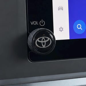 May include: A black circular volume control knob with a textured knurled edge and the Toyota logo centered on the face. The knob is mounted on a black dashboard panel next to a digital screen displaying a volume icon, a car icon, and a search icon.