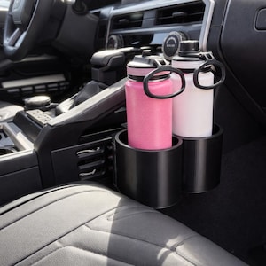 May include: A black car cup holder with two insulated water bottles. One bottle is pink, the other is white. The bottles have black handles and are placed in a custom-fit holder inside a vehicle's interior.