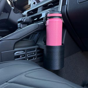 May include: A pink water bottle with a geometric pattern sits in a black cup holder inside a car. The cup holder is mounted on the center console, near the dashboard and passenger seat.