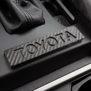 May include: A rectangular black carbon fiber inlay featuring the embossed text TOYOTA in a bold font. The inlay is positioned on a matte black textured plastic surface within a vehicle interior, near a black leather gear shift boot.