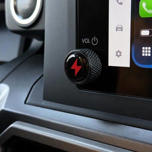 May include: A black circular volume control knob with a textured knurled edge and a glossy center featuring a red lightning bolt icon. The knob is attached to the side of a vehicle's infotainment screen, which displays the text VOL and a power symbol.