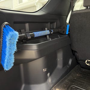 May include: A blue and black car wash brush and squeegee combo, stored in a vehicle's interior. The brush has blue bristles, and the squeegee has a blue blade. The handle is black with blue and yellow accents.