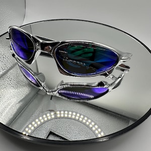 Op de afbeelding: Zilverkleurige zonnebril met blauwe glazen op een reflecterend oppervlak. De zonnebril heeft een strak, modern design. De glazen reflecteren de omgeving, waardoor een levendig, veelkleurig effect ontstaat. De afbeelding is goed belicht en benadrukt de details.