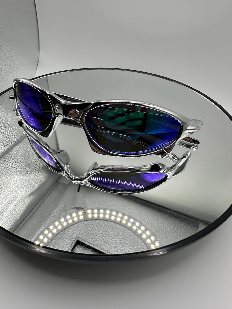 Op de afbeelding: Zilverkleurige zonnebril met blauwe glazen op een reflecterend oppervlak. De zonnebril heeft een strak, modern design. De glazen reflecteren de omgeving, waardoor een levendig, veelkleurig effect ontstaat. De afbeelding is goed belicht en benadrukt de details.