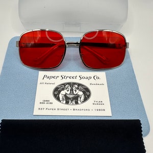 May include: Silver framed square sunglasses with red lenses. The sunglasses are on a blue background with a white card that reads "Paper Street Soap Co. All Natural Handmade (288) 555-0153 TYLER DURDEN 537 PAPER STREET BRADFORD 19808"