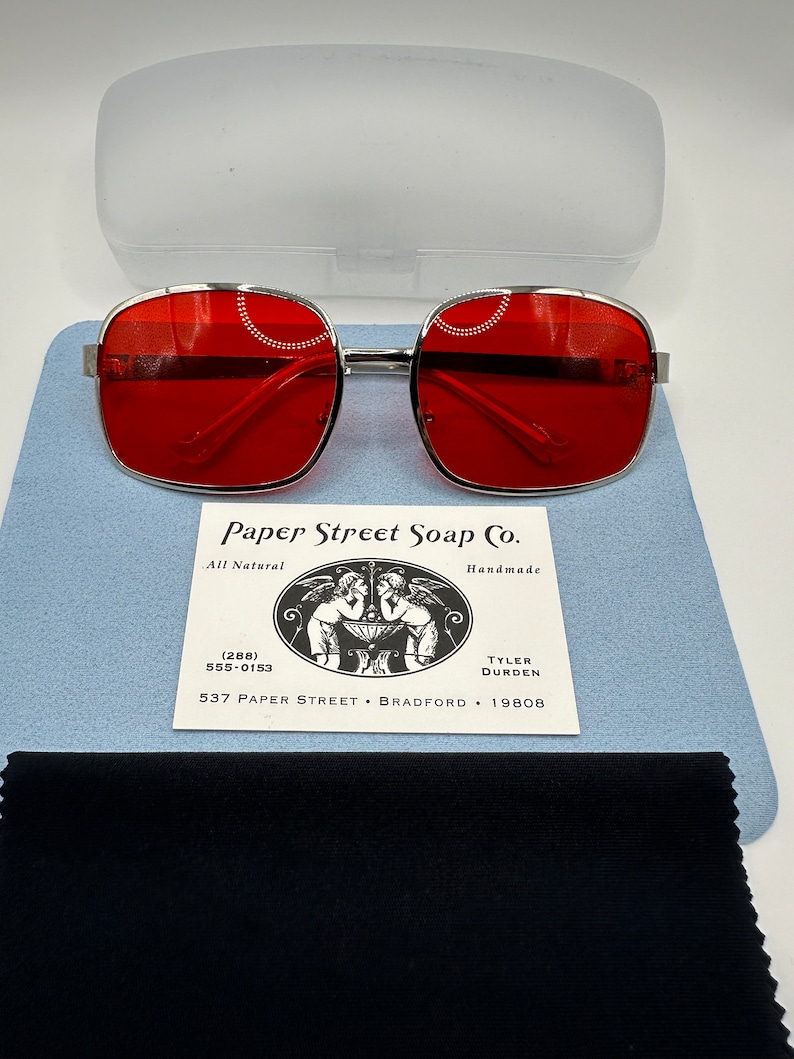 May include: Silver framed square sunglasses with red lenses. The sunglasses are on a blue background with a white card that reads "Paper Street Soap Co. All Natural Handmade (288) 555-0153 TYLER DURDEN 537 PAPER STREET BRADFORD 19808"