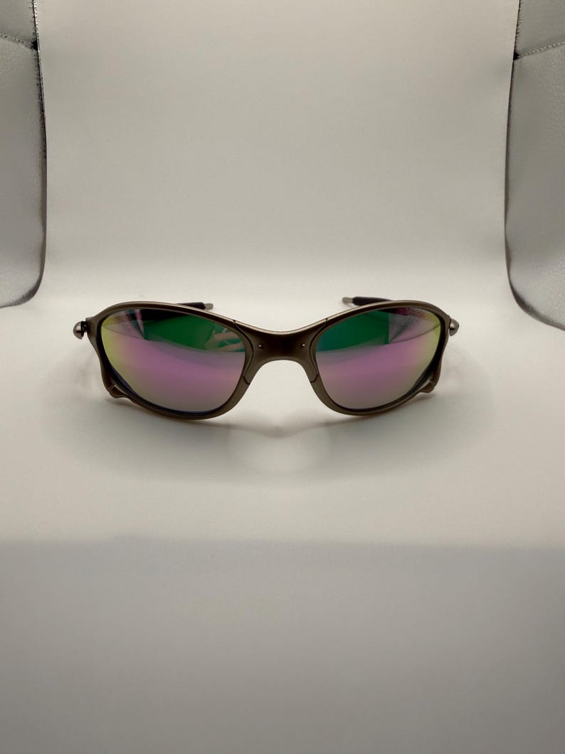 May include: A pair of sunglasses with a dark brown frame and iridescent lenses that reflect shades of pink, green, and purple. The sunglasses have a curved design and are set against a white background.