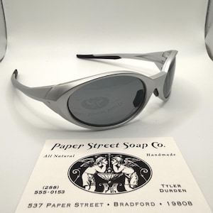 Op de afbeelding: Zilverkleurige zonnebril met donkere glazen. De zonnebril heeft een modern design. Een visitekaartje met de tekst "Paper Street Soap Co." ligt eronder.