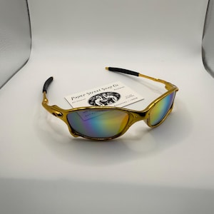 May include: Gold-framed sunglasses with iridescent lenses. The sunglasses have a sleek, modern design with a gold finish. The lenses reflect a rainbow of colors. A small card with the text "Paper Street Soap Co." is visible.