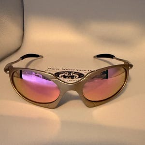 May include: Gold-tone sunglasses with pink mirrored lenses. The glasses have a sleek, modern design with a curved frame and black accents on the temples. The text "Paper Street Soap Co." is visible above the glasses.