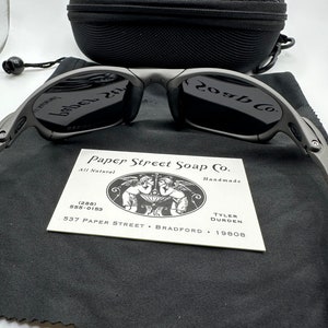 May include: A pair of black sunglasses with a gray frame and a black case. The sunglasses have a logo that says "20gb Co." on the right lens and "babel 2.1" on the left lens. The case has a zipper and a black drawstring bag with the text "Paper Street Soap Co. All Natural Handmade (288) 555-0153 537 PAPER STREET BRADFORD 19808 TYLER DURDEN".