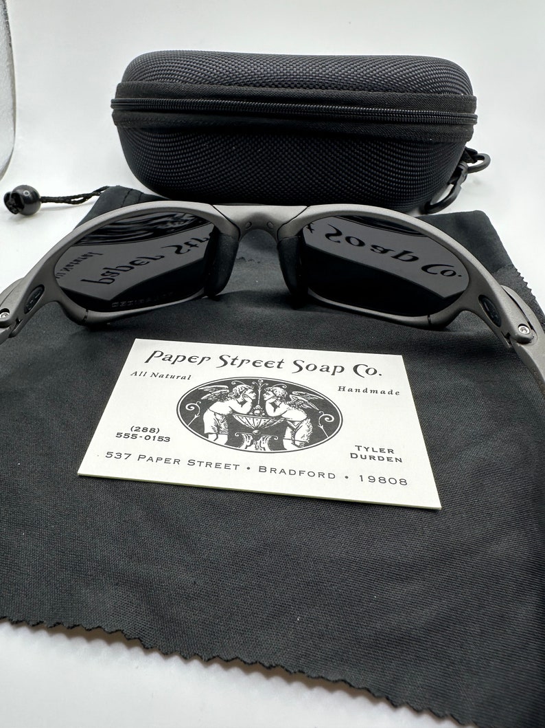 May include: A pair of black sunglasses with a gray frame and a black case. The sunglasses have a logo that says "20gb Co." on the right lens and "babel 2.1" on the left lens. The case has a zipper and a black drawstring bag with the text "Paper Street Soap Co. All Natural Handmade (288) 555-0153 537 PAPER STREET BRADFORD 19808 TYLER DURDEN".