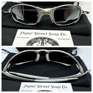 May include: Silver-framed sunglasses with dark lenses, resting on a black cloth. The glasses have a distinctive, angular design. A card with the text "Paper Street Soap Co." is visible.