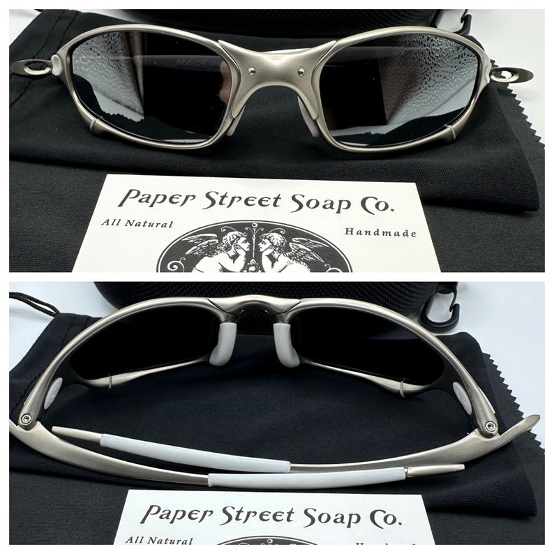 May include: Silver-framed sunglasses with dark lenses, resting on a black cloth. The glasses have a distinctive, angular design. A card with the text "Paper Street Soap Co." is visible.
