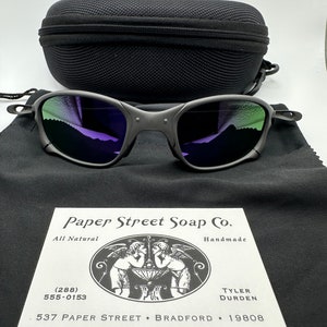 May include: A black and silver pair of sunglasses with a purple and green mirrored lens. The sunglasses are in a black case with a business card for Paper Street Soap Co. The business card has a black and white illustration of two angels and the text "All Natural", "Handmade", "(288) 555-0153", "537 Paper Street Bradford 19808", "Tyler Durden".