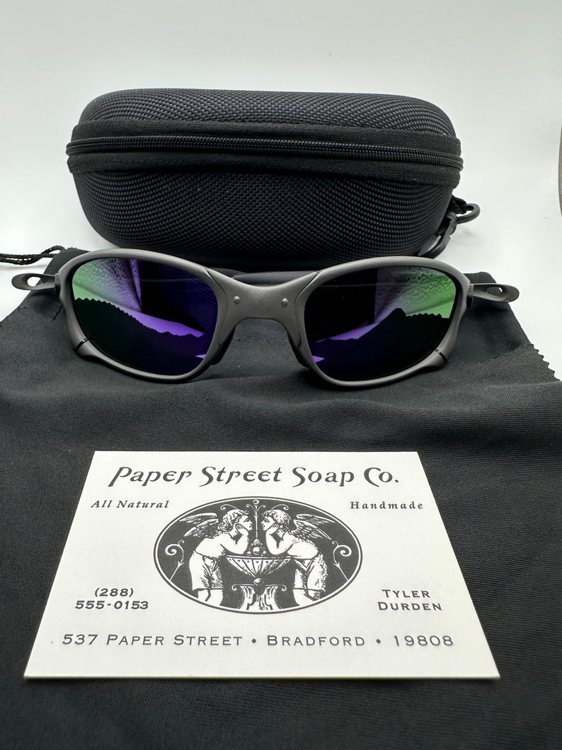 May include: A black and silver pair of sunglasses with a purple and green mirrored lens. The sunglasses are in a black case with a business card for Paper Street Soap Co. The business card has a black and white illustration of two angels and the text "All Natural", "Handmade", "(288) 555-0153", "537 Paper Street Bradford 19808", "Tyler Durden".